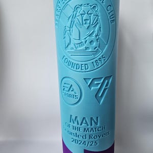 Op de afbeelding: Een blauwe en paarse plastic trofee met de tekst "Bearsted Football Club Founded 1895" en "Man of the Match Bearsted Rovers 2024/25". De trofee heeft een logo van een beer met een voetbal.