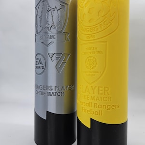 May include: Two silver and yellow trophy cups with black bases. The silver cup has a logo with trees and the text "SILVARC" and "EA SPORTS". The yellow cup has a logo with a soccer ball and the text "KOMERSALL RANGERS F.C. 1961". Both cups have the text "MANAGERS PLAYER OF THE MATCH" and "PLAYER OF THE MATCH".