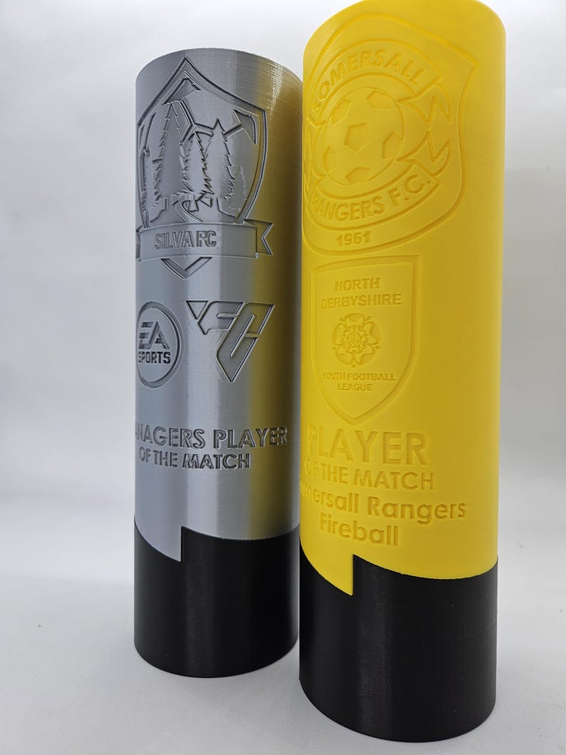May include: Two silver and yellow trophy cups with black bases. The silver cup has a logo with trees and the text "SILVARC" and "EA SPORTS". The yellow cup has a logo with a soccer ball and the text "KOMERSALL RANGERS F.C. 1961". Both cups have the text "MANAGERS PLAYER OF THE MATCH" and "PLAYER OF THE MATCH".
