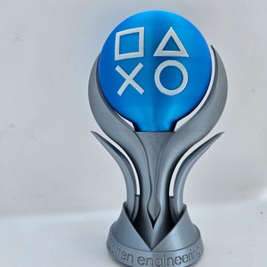 May include: A silver 3D printed trophy with a blue sphere on top. The sphere has the PlayStation button symbols in white. The base of the trophy has the words "otten engineering" inscribed on it.