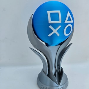 Puede incluir: Un trofeo plateado con una esfera azul en la parte superior. La esfera tiene los símbolos de los botones de PlayStation. La base del trofeo dice "Cotten engineer".
