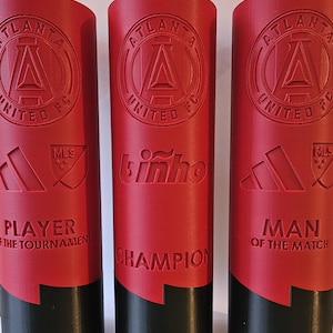 May include: Three red and black trophy cups with the Atlanta United FC logo, the words "Player of the Tournament", "Champion", and "Man of the Match", and the MLS logo.
