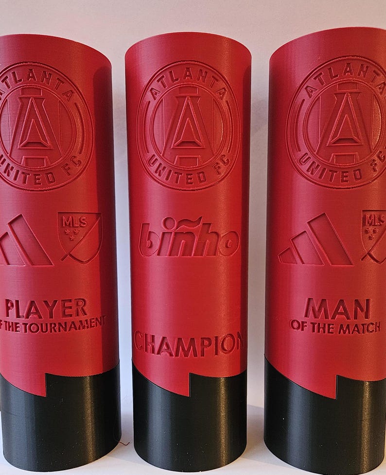 May include: Three red and black trophy cups with the Atlanta United FC logo, the words "Player of the Tournament", "Champion", and "Man of the Match", and the MLS logo.