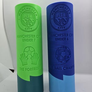 May include: Two green and blue plastic trophies with the Manchester City Football Club logo. The green trophy says "Manchester City Under 7 The Fortress" and the blue trophy says "Manchester City Under 6 Five Ball Champions".