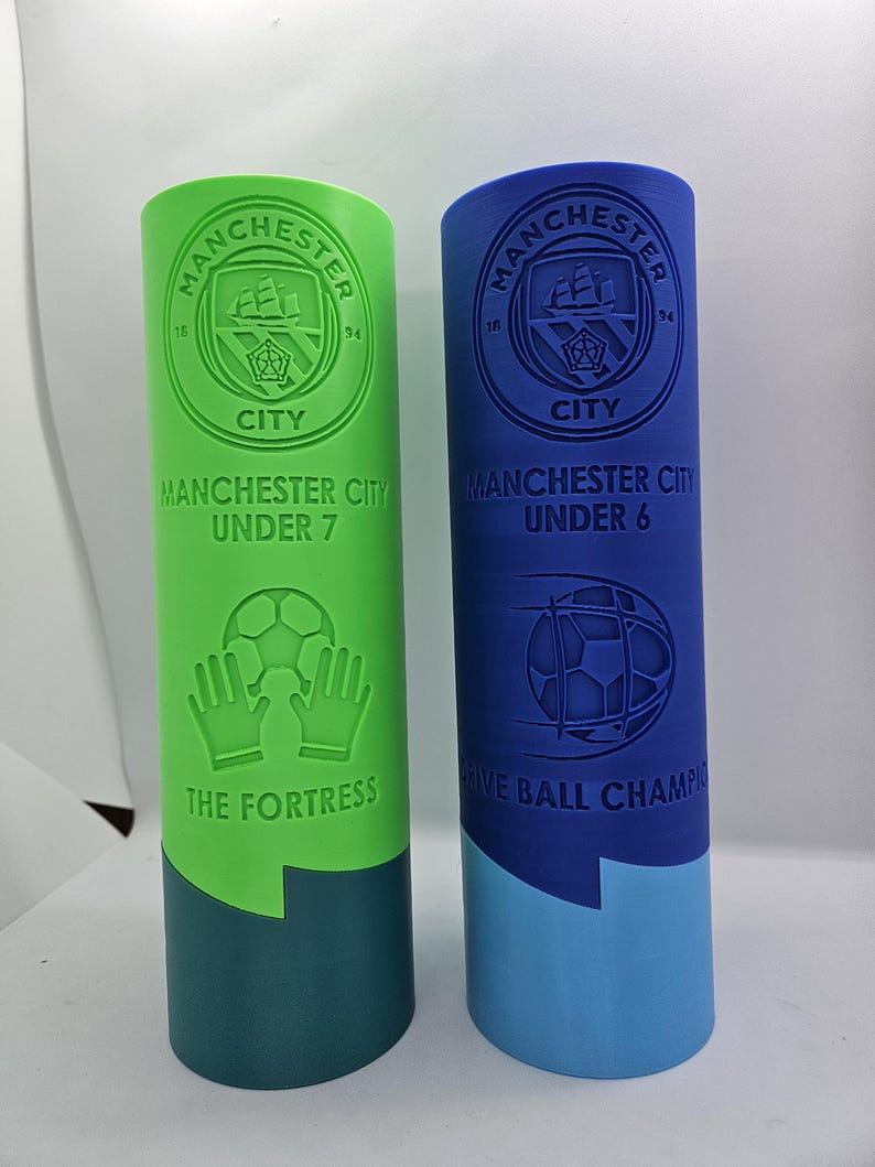 May include: Two green and blue plastic trophies with the Manchester City Football Club logo. The green trophy says "Manchester City Under 7 The Fortress" and the blue trophy says "Manchester City Under 6 Five Ball Champions".