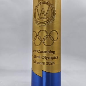 May include: A gold trophy with a blue base. The trophy has the text "AW Coaching Football Olympics Winners 2024" and a logo with a crown and the letters "AW" inside a circle.