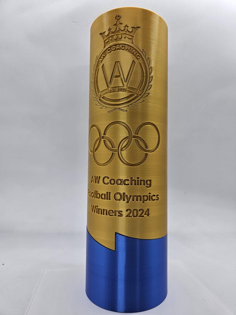 May include: A gold trophy with a blue base. The trophy has the text "AW Coaching Football Olympics Winners 2024" and a logo with a crown and the letters "AW" inside a circle.