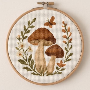Könnte beinhalten: Kreuzstich-Stickerei mit zwei braunen Pilzen, einem Schmetterling und Blättern in einem Holzreifen. Die Pilze haben beige Stiele und braune Kappen. Die Blätter sind grün und orange, und der Schmetterling ist braun. Der Hintergrund ist cremefarben.