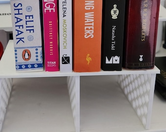 May include: A collection of hardcover books with colorful spines, including titles like "There Are Rivers in the Sky" and "The Hobbit." The books are arranged on a white shelf, showcasing a variety of literary works.