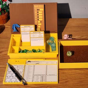 Peut inclure: Un ensemble de jeu de rôle de table avec une boîte marron et jaune, des dés, des fiches de personnage et des figurines miniatures. L'ensemble comprend un stylo, des cartes et un bac à dés. La boîte a un indicateur d'emplacement de sorts.