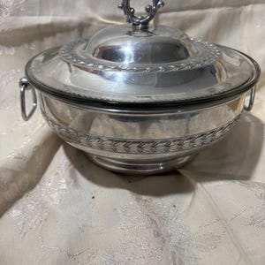 May include: A silver-colored serving bowl with a lid. The bowl has a decorative pattern around the rim and base. The lid features a handle with a circular design. The bowl has two handles on the sides.