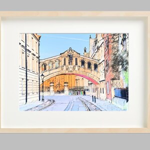 Può includere: Stampa artistica incorniciata di una scena di strada con un ponte ed edifici. L'opera d'arte è in stile acquerello, presentata in una cornice di legno chiaro con passe-partout bianco. Il ponte ha un design ad arco.