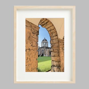 Può includere: Stampa artistica incorniciata con un'illustrazione in stile schizzo di un edificio con cupola, visto attraverso un arco. L'arco è in pietra e il cielo è blu. La cornice è in legno chiaro.