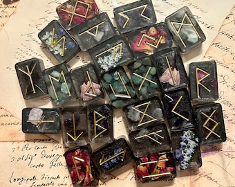 May include: A set of 18 black and gold rune stones with various floral and crystal accents. The runes are set in clear resin and are arranged on a piece of old paper.