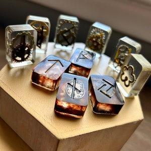 May include: A set of eight clear resin dice with various symbols and gears embedded inside. The dice are arranged in a circle on a wooden hexagonal stand.