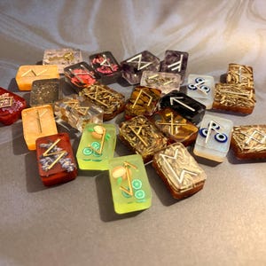 May include: A collection of rectangular resin tiles featuring embedded decorative elements such as dried flowers, glitter, fruit slices, and evil eye beads. Each tile is topped with a metallic gold-colored rune symbol, creating a diverse set of small, polished, and translucent pieces with varied textures and colors.