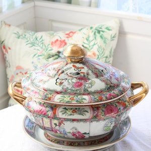 May include: A decorative porcelain soup tureen with a lid, handles, and a matching plate. The tureen is white with colorful floral and bird designs, accented with gold trim. The lid has a gold knob.