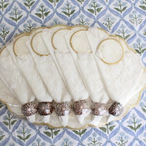 May include: A set of white cloth napkins with decorative floral embroidery, each held by a brown and white patterned napkin ring. The napkins are arranged on a scalloped, cream-colored plate with a gold trim. The background is a blue and white floral patterned fabric.