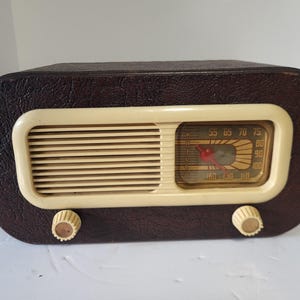 Könnte beinhalten: Ein Vintage-Radio der Marke Philco mit einem dunkelbraunen, texturierten Gehäuse und einer cremefarbenen Frontblende. Das Radio verfügt über ein horizontales Lautsprechergitter, eine rechteckige Skala mit rotem Zeiger und zwei cremefarbene Drehknöpfe.
