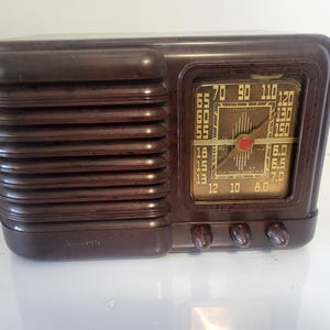 May include: A vintage brown radio with a rectangular design. The radio features a speaker grill on the left side and a dial with numbers and a needle on the right. The radio has three knobs at the bottom.