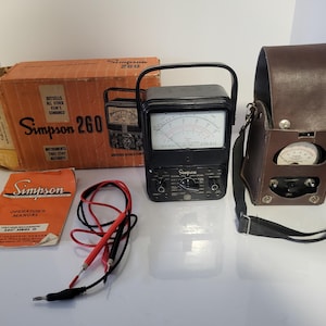 May include: A vintage Simpson 260 multimeter set. The set includes a black multimeter with a handle, a brown leather carrying case, a red and black test lead, an orange manual, and a cardboard box with the Simpson 260 logo.