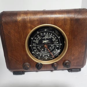 May include: A brown wooden radio with a large circular black dial featuring white numbers and the Zenith logo in the center. The radio has a gold-toned bezel around the dial and four dark brown control knobs along the bottom front edge.