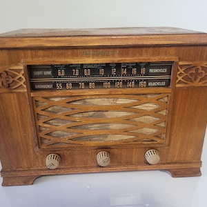 May include: A vintage wooden radio with a dark finish. The radio features a speaker grill, tuning dials, and a display with frequency markings. The design includes decorative carvings on the sides. The radio's display shows "SHORT WAVE" and "BROADCAST" frequencies.