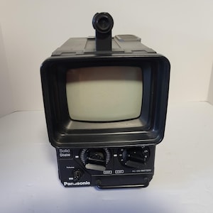 Puede incluir: Un televisor portátil Panasonic vintage con carcasa de plástico negro y una pantalla pequeña. El panel frontal presenta dos grandes perillas de sintonización circulares, un dial de volumen más pequeño y el texto Solid State, UHF, VHF, AC/DC/BATTERY, Panasonic.