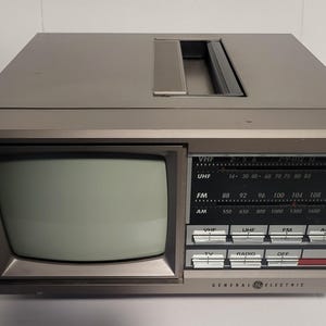 Puede incluir: Un combo de radio y televisión portátil General Electric vintage. El dispositivo gris plateado presenta una pequeña pantalla, un dial de radio con frecuencias AM/FM y botones de control. La parte superior tiene un asa.