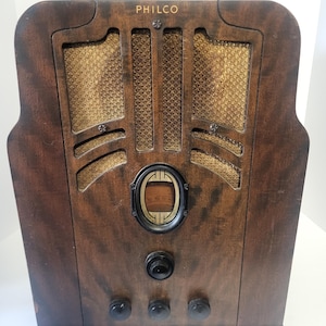 May include: A vintage Philco tabletop radio featuring a dark brown wooden cabinet with a vertical grain pattern. The front includes a gold-colored fabric speaker grille, a central oval-shaped tuning dial, and four black circular control knobs.