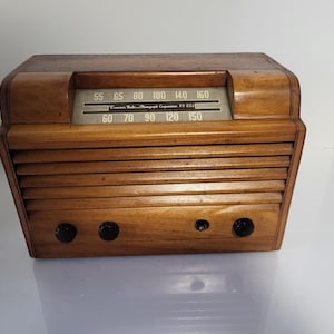 May include: A vintage wooden radio in a light brown finish. The radio has a horizontal speaker grill, a tuning dial with numbers, and three black knobs. The text "Emerson Radio & Phonograph Corporation" is visible on the dial.