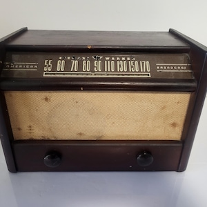 Pode incluir: Um rádio de madeira castanho-escuro com uma grelha de altifalante em tecido bege. O painel superior apresenta o texto Stewart-Warner, American e Broadcast, juntamente com um mostrador de frequência com números de 55 a 170. Dois botões pretos.