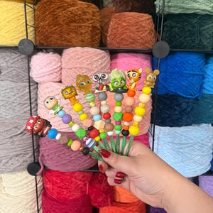 Puede incluir: Una colecci&oacute;n de adornos coloridos para ganchos de ganchillo con dise&ntilde;os de personajes de dibujos animados. Los ganchos se sostienen frente a un fondo de madejas de hilo de varios colores, incluyendo rosa, marr&oacute;n y azul.