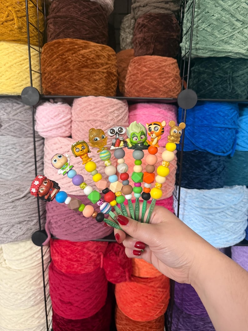 Puede incluir: Una colecci&oacute;n de adornos coloridos para ganchos de ganchillo con dise&ntilde;os de personajes de dibujos animados. Los ganchos se sostienen frente a un fondo de madejas de hilo de varios colores, incluyendo rosa, marr&oacute;n y azul.