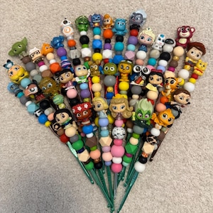 Puede incluir: Una colecci&oacute;n de ganchos de ganchillo con cuentas de colores y figuras de personajes. Los ganchos son verdes y las cuentas son de varios colores, incluyendo rosa, azul, amarillo y negro. Las figuras son de personajes de dibujos animados populares.