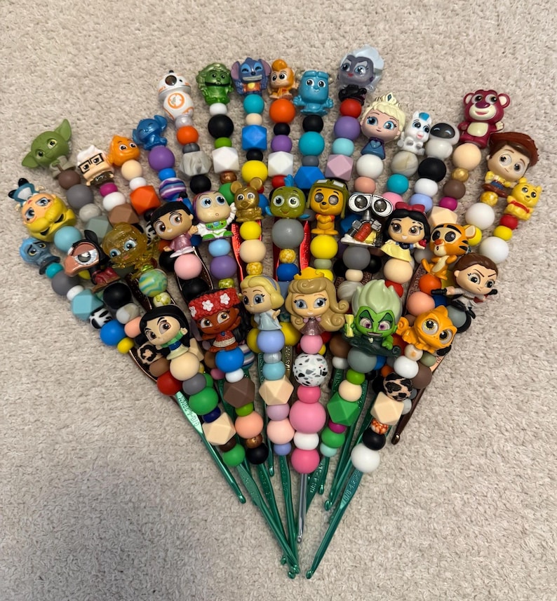 Puede incluir: Una colecci&oacute;n de ganchos de ganchillo con cuentas de colores y figuras de personajes. Los ganchos son verdes y las cuentas son de varios colores, incluyendo rosa, azul, amarillo y negro. Las figuras son de personajes de dibujos animados populares.