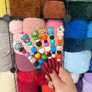 Puede incluir: Un conjunto de ganchillos rojos con adornos de personajes y cuentas coloridas. Cada ganchillo presenta un personaje diferente y un dise&ntilde;o &uacute;nico. El fondo muestra madejas de hilo de varios colores, incluyendo rosa, marr&oacute;n y azul.