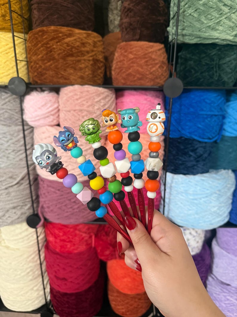 Puede incluir: Un conjunto de ganchillos rojos con adornos de personajes y cuentas coloridas. Cada ganchillo presenta un personaje diferente y un dise&ntilde;o &uacute;nico. El fondo muestra madejas de hilo de varios colores, incluyendo rosa, marr&oacute;n y azul.