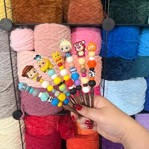 Puede incluir: Una mano sostiene varios ganchillos con adornos de cuentas tem&aacute;ticas de personajes. Los ganchillos tienen mangos marrones y est&aacute;n adornados con cuentas de colores y figuritas de personajes. El fondo presenta estantes llenos de varios colores de hilo.