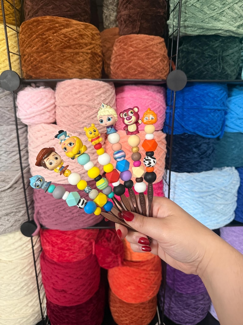 Puede incluir: Una mano sostiene varios ganchillos con adornos de cuentas tem&aacute;ticas de personajes. Los ganchillos tienen mangos marrones y est&aacute;n adornados con cuentas de colores y figuritas de personajes. El fondo presenta estantes llenos de varios colores de hilo.