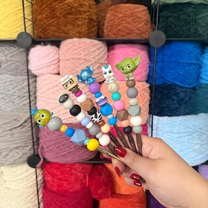Puede incluir: Ganchos de ganchillo con adornos de personajes, como Yoda y Stitch. Los ganchos tienen cuentas decorativas y se presentan frente a un fondo de madejas de hilo de colores. Ideal para tejer.