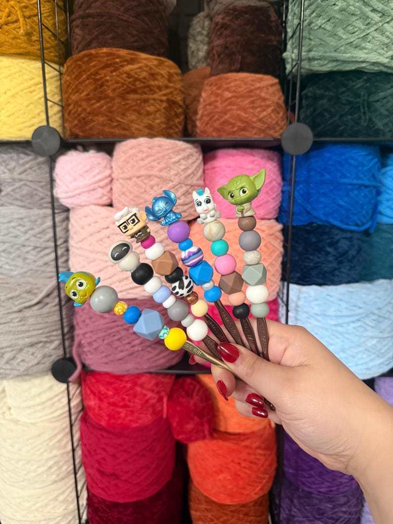 Puede incluir: Ganchos de ganchillo con adornos de personajes, como Yoda y Stitch. Los ganchos tienen cuentas decorativas y se presentan frente a un fondo de madejas de hilo de colores. Ideal para tejer.