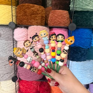 Puede incluir: Una colecci&oacute;n de ganchillos de ganchillo coloridos con adornos tem&aacute;ticos de personajes, que incluyen figuras de dibujos animados y dise&ntilde;os de animales. Los ganchos presentan varias combinaciones de cuentas en tonos rosa, negro y blanco, sobre un fondo de madejas de hilo.