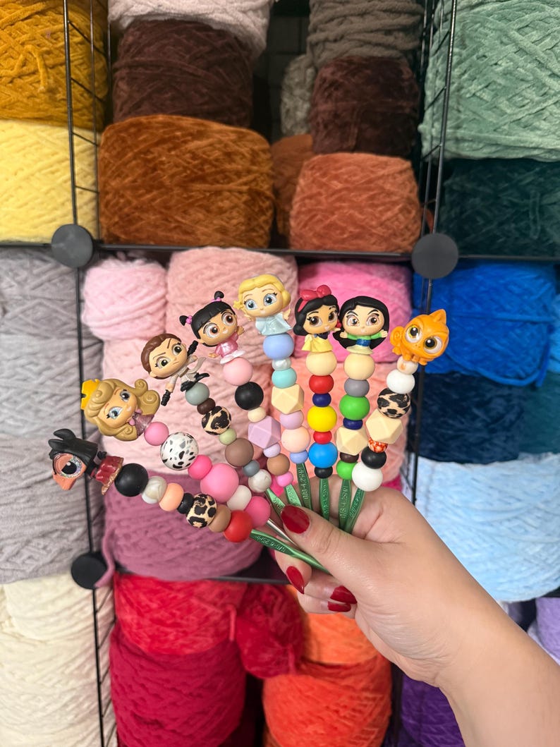 Puede incluir: Una colecci&oacute;n de ganchillos de ganchillo coloridos con adornos tem&aacute;ticos de personajes, que incluyen figuras de dibujos animados y dise&ntilde;os de animales. Los ganchos presentan varias combinaciones de cuentas en tonos rosa, negro y blanco, sobre un fondo de madejas de hilo.