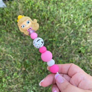 Puede incluir: Bol&iacute;grafo de cuentas con una figura de princesa de dibujos animados con una corona dorada. El bol&iacute;grafo tiene cuentas rosas, blancas y moteadas, y una varilla de metal plateado. El bol&iacute;grafo se sostiene sobre un fondo de hierba verde.