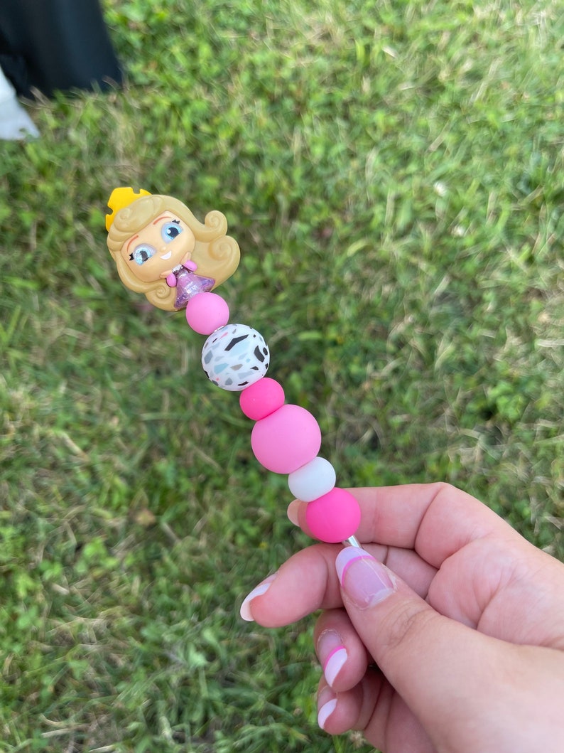 Puede incluir: Bol&iacute;grafo de cuentas con una figura de princesa de dibujos animados con una corona dorada. El bol&iacute;grafo tiene cuentas rosas, blancas y moteadas, y una varilla de metal plateado. El bol&iacute;grafo se sostiene sobre un fondo de hierba verde.