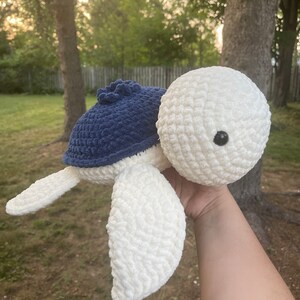 May include: A crocheted plush toy turtle with a white body and blue shell. The turtle has a small black eye and is being held by a hand.