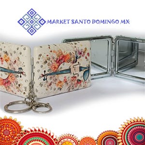 May include: A white, folding compact mirror with a floral and violin design. The mirror has a silver-colored frame and a key ring attachment. The text "MARKET SANTO DOMINGO MX" is visible in the background.