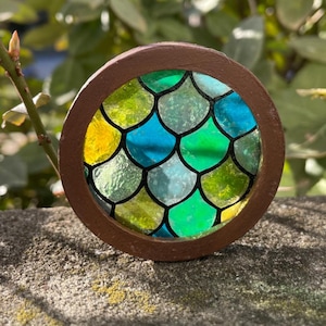 Può includere: Un oggetto rotondo in vetro colorato con una cornice marrone. Il vetro presenta un motivo a scaglie nei toni del giallo, verde e blu. L'oggetto è posto su uno sfondo verde sfocato, forse all'aperto.