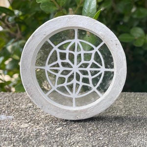 May include: A white circular decorative piece with a round window featuring a white geometric design. The window is made of clear glass with a frosted effect.
